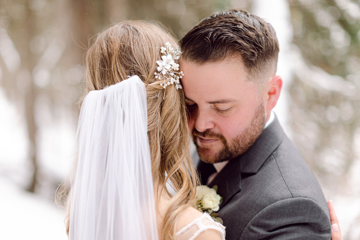 A couple shares a tender embrace, showcasing the bride's elegant hairstyle adorned with a floral hairpiece and her flowing veil, against a snowy backdrop.