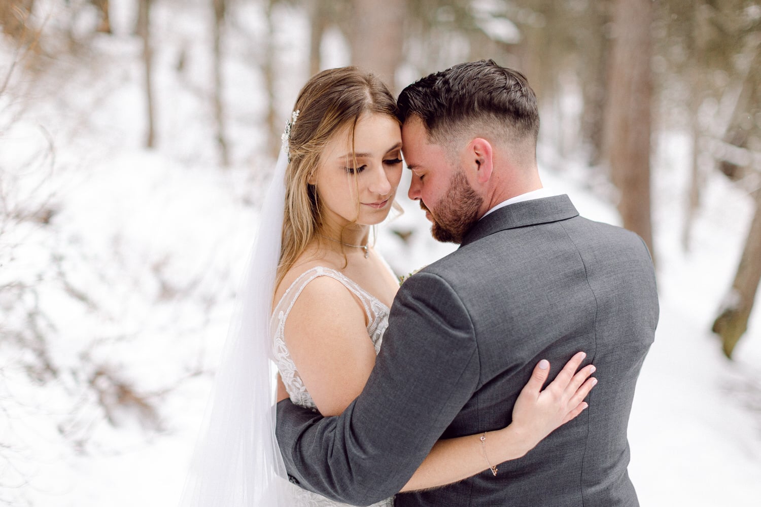 A couple shares a tender moment in a snowy forest, with the bride in a delicate gown and veil, and the groom in a stylish gray suit.