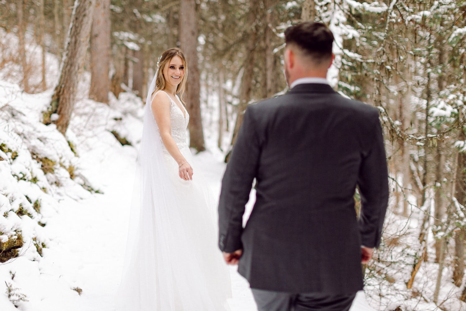 Bride in a snowy forest turning to smile at her groom in a dark suit.