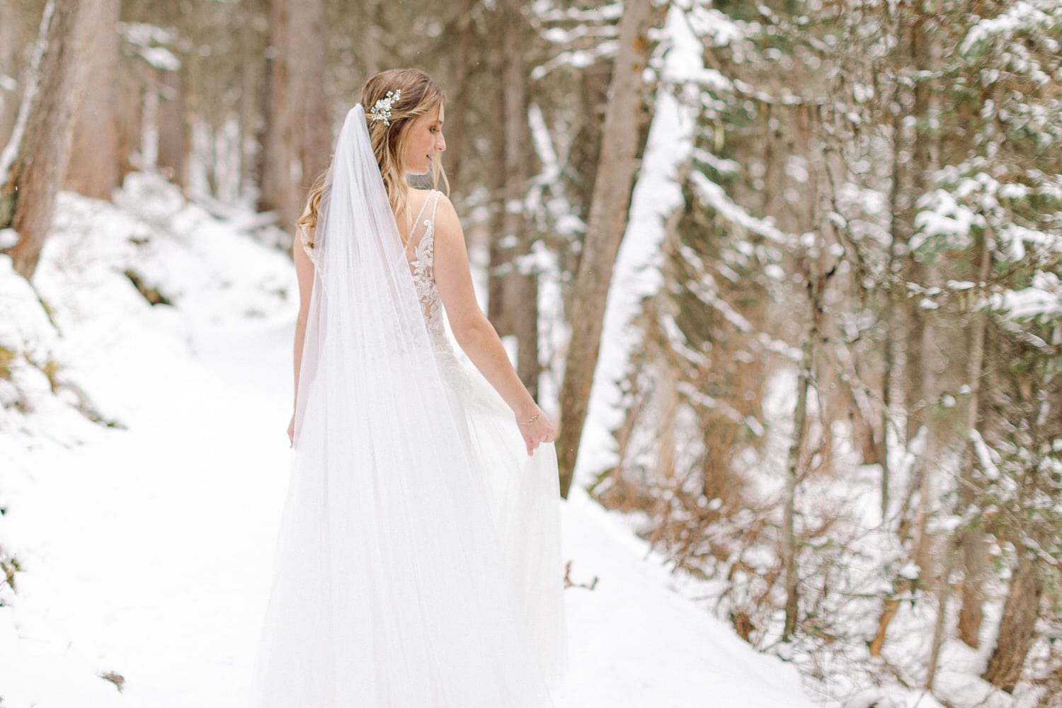 A bride in a flowing white gown and veil walks through a snow-covered forest, surrounded by tall trees.