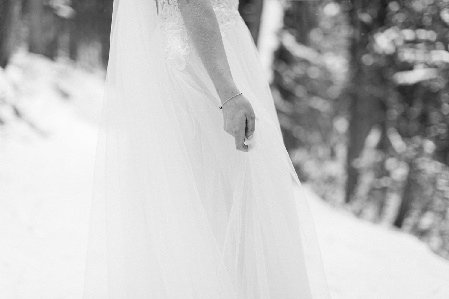 A close-up of a bride's hand draped in a flowing white dress, set against a snowy forest backdrop.