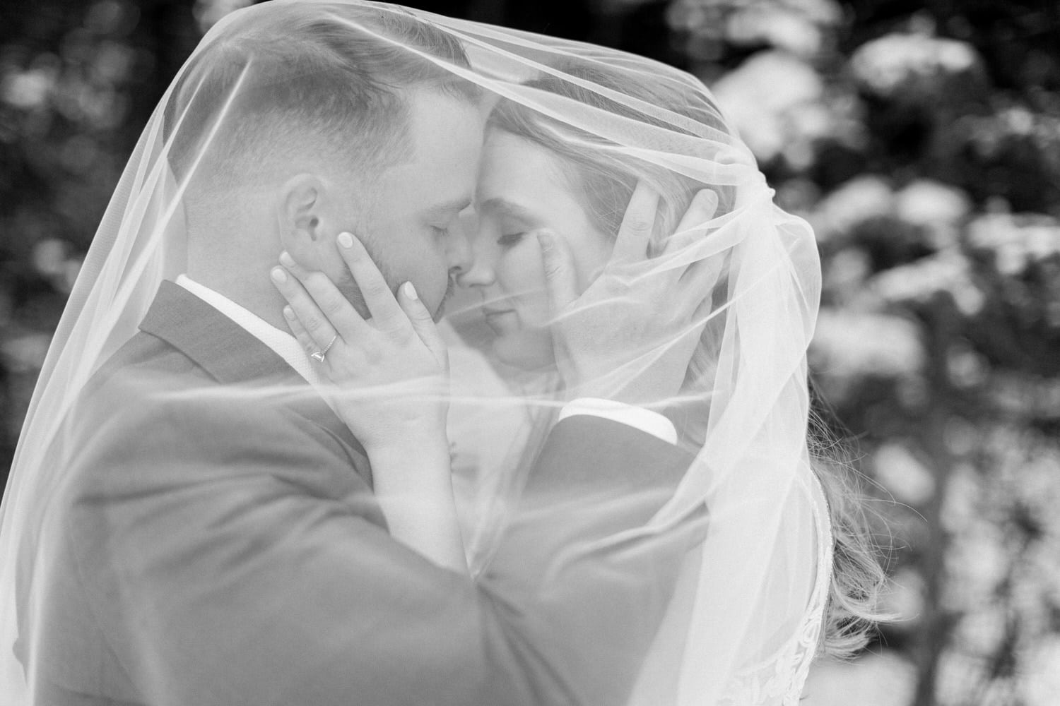 A couple shares a tender moment, their faces nearly touching, framed by a delicate veil in soft black and white.