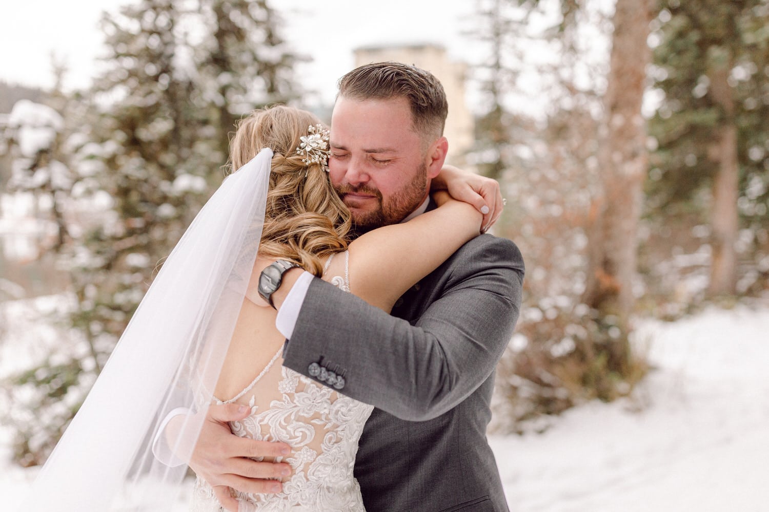 A groom tenderly embraces his bride, both emotional in a snowy outdoor setting, capturing a moment of love and connection.