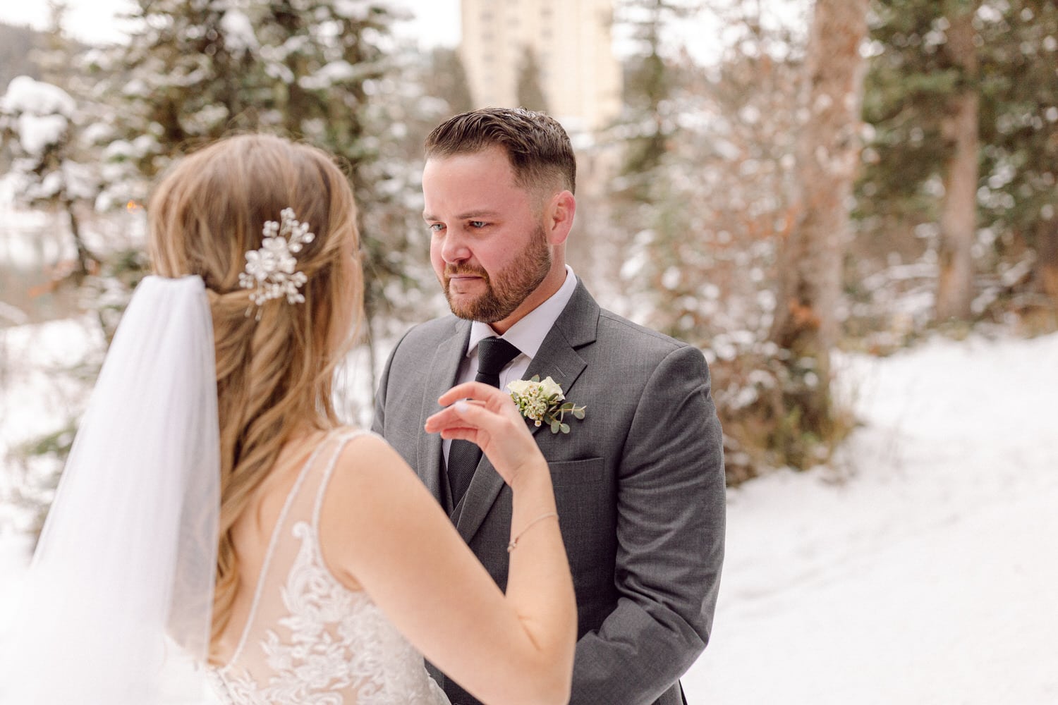 A bride and groom share an intimate moment outdoors in a snowy setting, surrounded by trees, as the bride gestures warmly while looking at the groom.