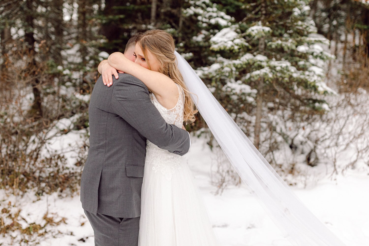 A bride and groom share a heartfelt hug in a snowy forest setting, capturing a moment of intimacy and love on their wedding day.