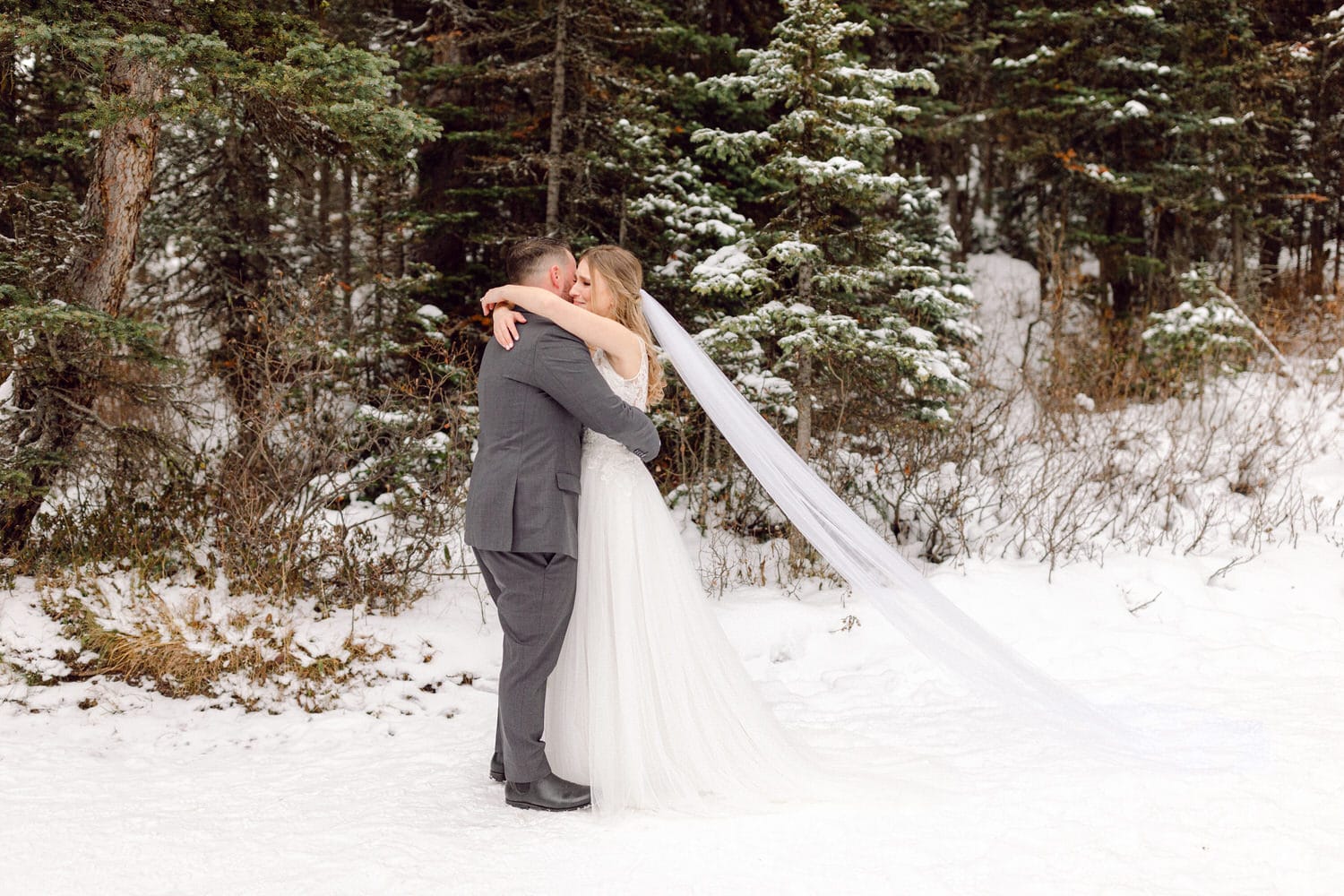 A couple shares a romantic hug in a snowy forest, with the bride in a flowing gown and veil, surrounded by evergreen trees.