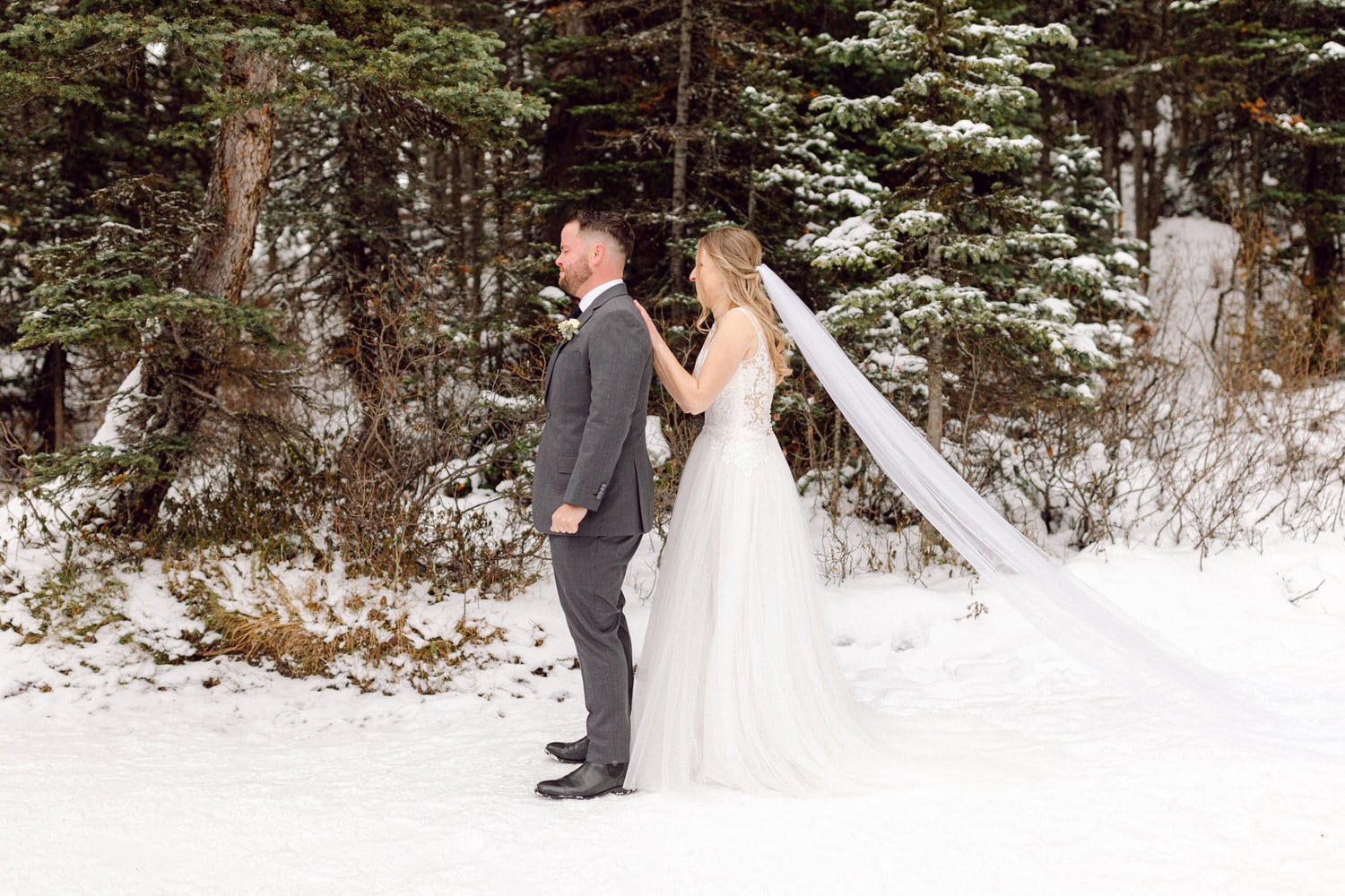 A bride and groom stand back-to-back in a snowy forest setting, with the bride adjusting her veil as the groom looks on.