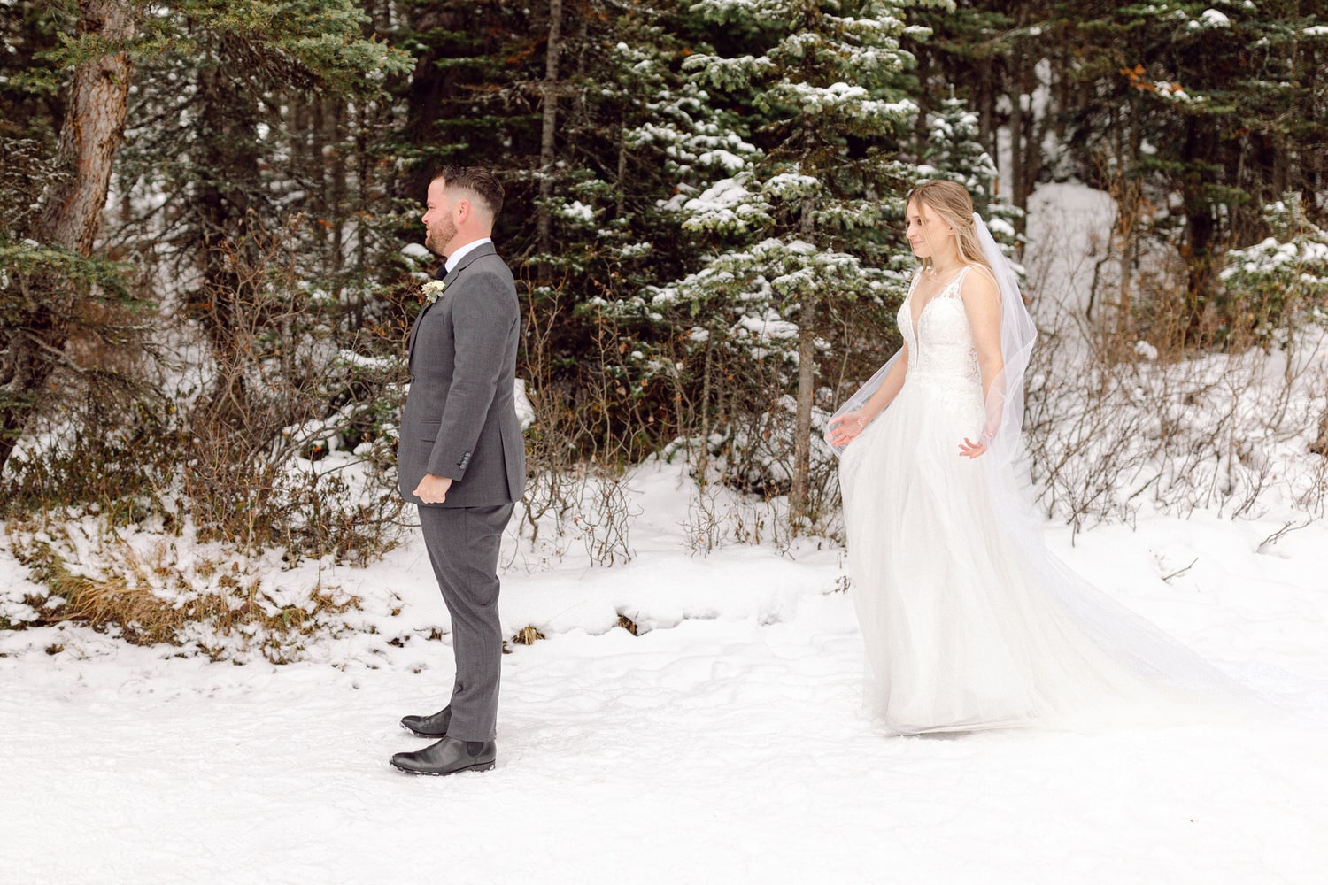 A groom and bride stand in a snowy forest, the bride approaching from behind as the groom faces away, showcasing a heartfelt wedding moment.