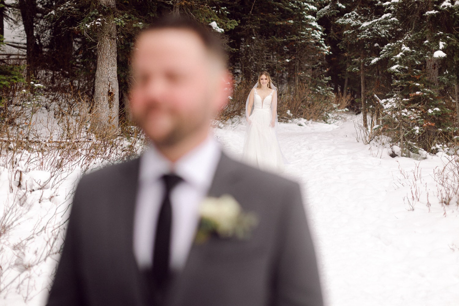 A bride walking down a snowy path while a blurred groom stands in the foreground, surrounded by trees.