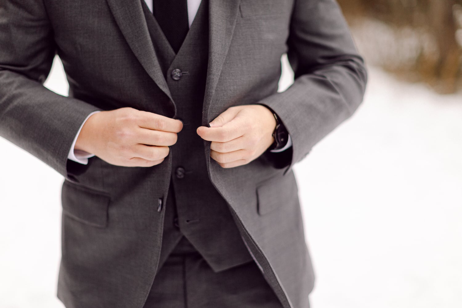 A man adjusting the buttons of his gray suit jacket, dressed smartly in a winter landscape.