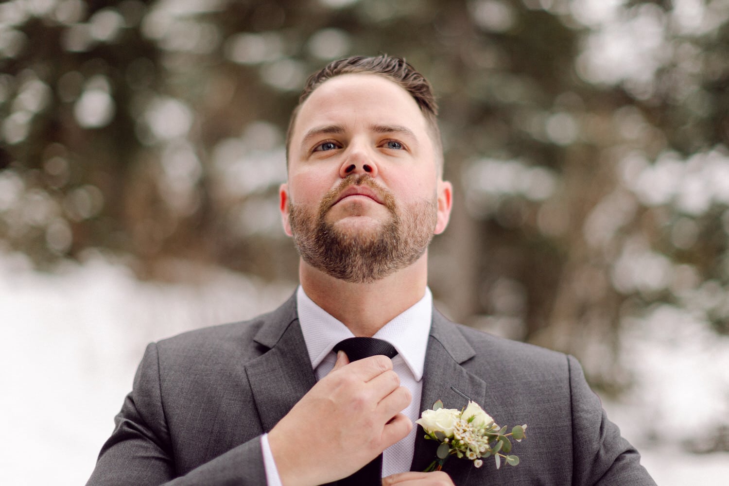 A well-dressed man adjusts his tie while wearing a gray suit and a white floral boutonniere against a blurred snowy background.