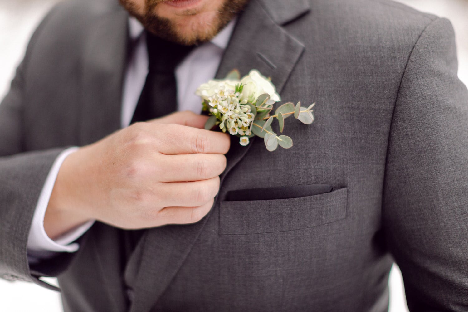 A close-up of a man in a gray suit adjusting a white floral boutonnière on his lapel, highlighting details of the attire and the flowers.