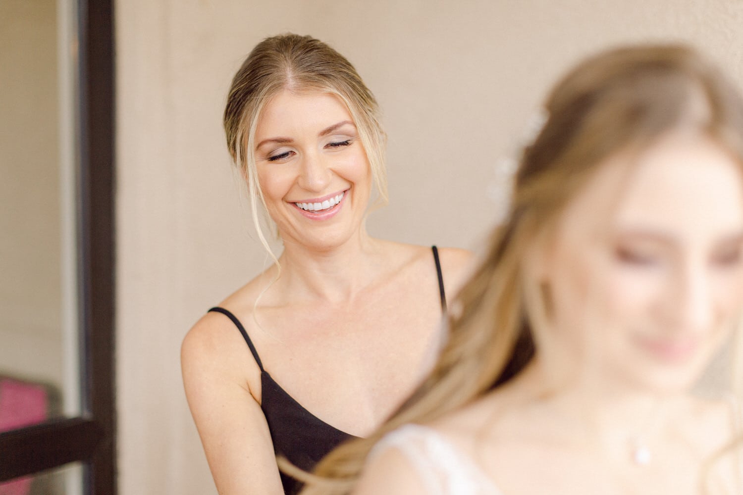 A smiling woman helps another woman prepare for a special occasion, showcasing friendship and elegance.