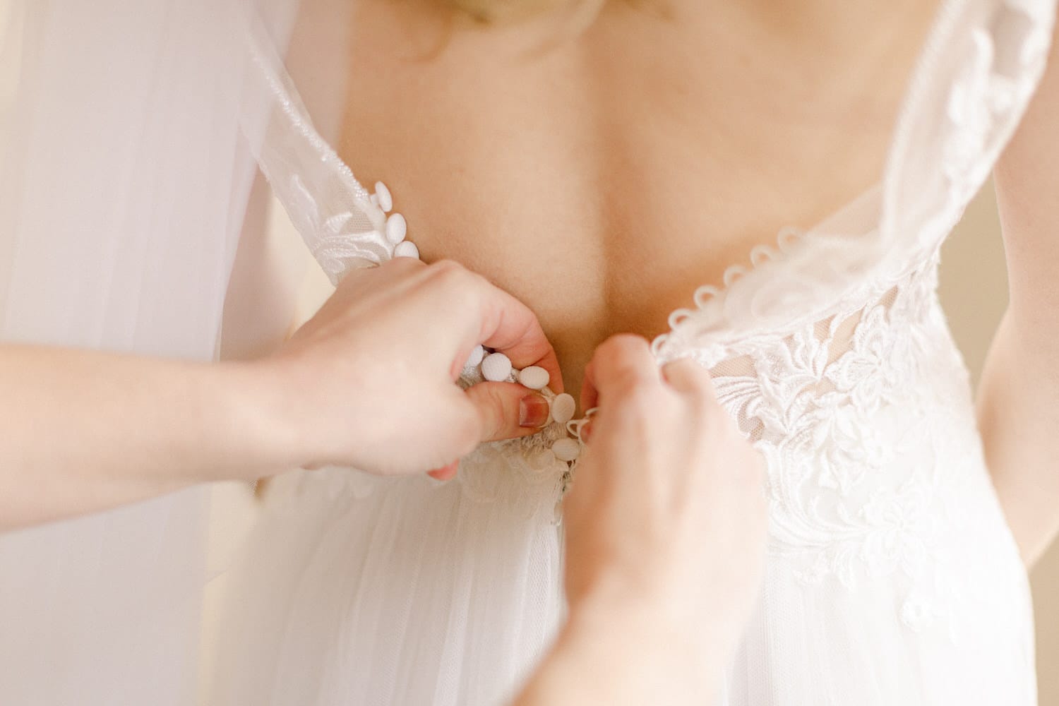 A close-up of hands fastening buttons on the back of a wedding dress adorned with intricate lace details.