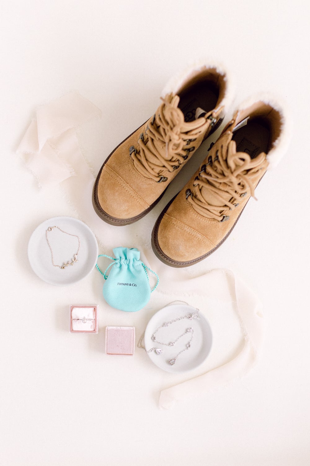 A pair of beige winter boots surrounded by elegant jewelry pieces and a Tiffany & Co. pouch on a soft background.