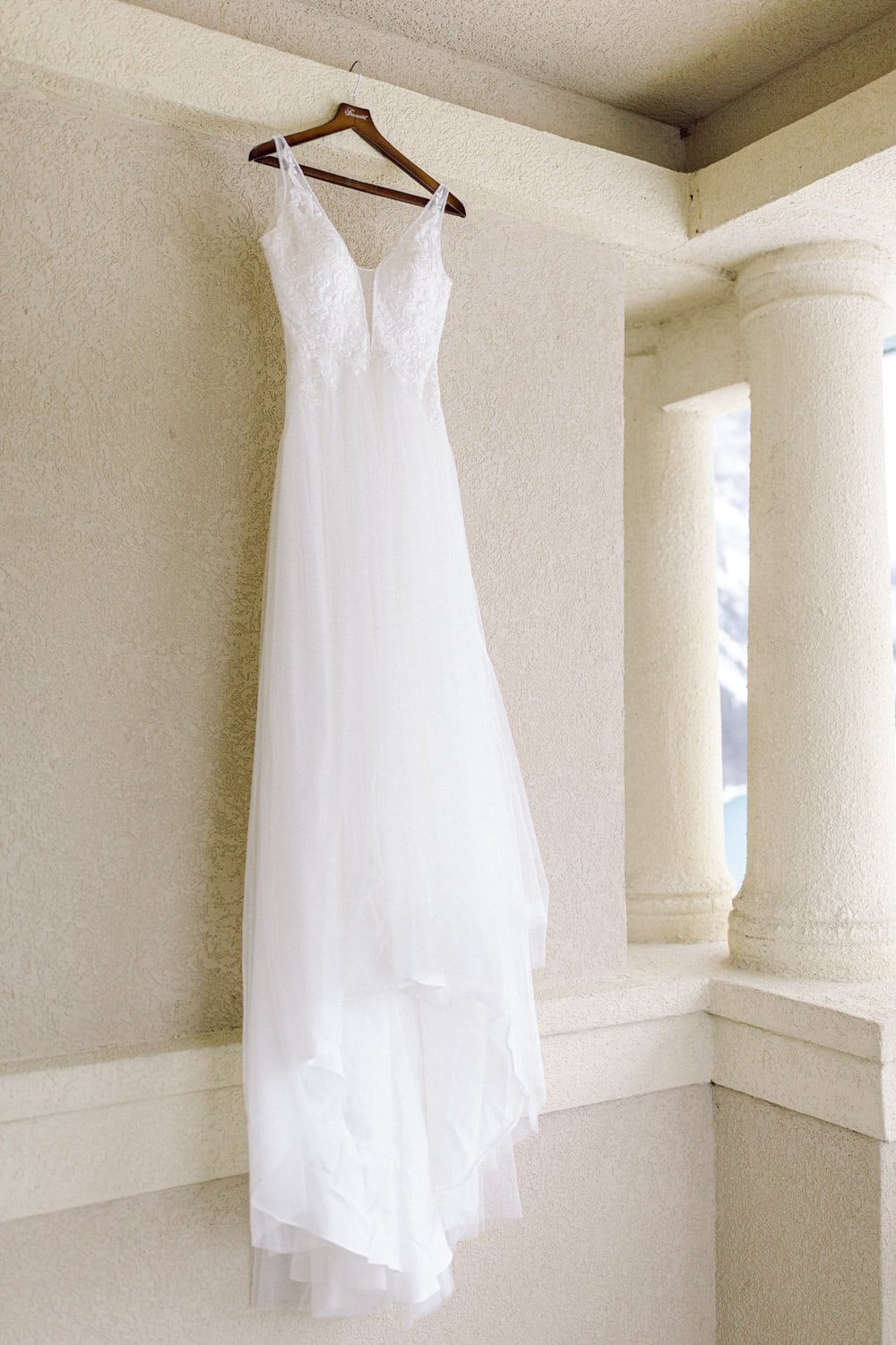 A delicate white wedding dress with lace detailing hangs on a wooden hanger against a textured wall, showcasing its flowing silhouette.