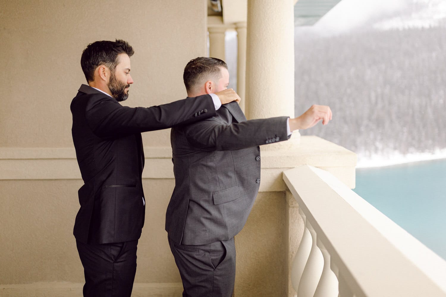 Two men in suits adjusting each other's attire on a balcony overlooking a scenic view.