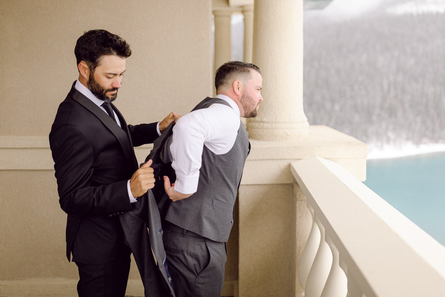 Two men in formal attire adjusting suits on a balcony with a scenic backdrop.