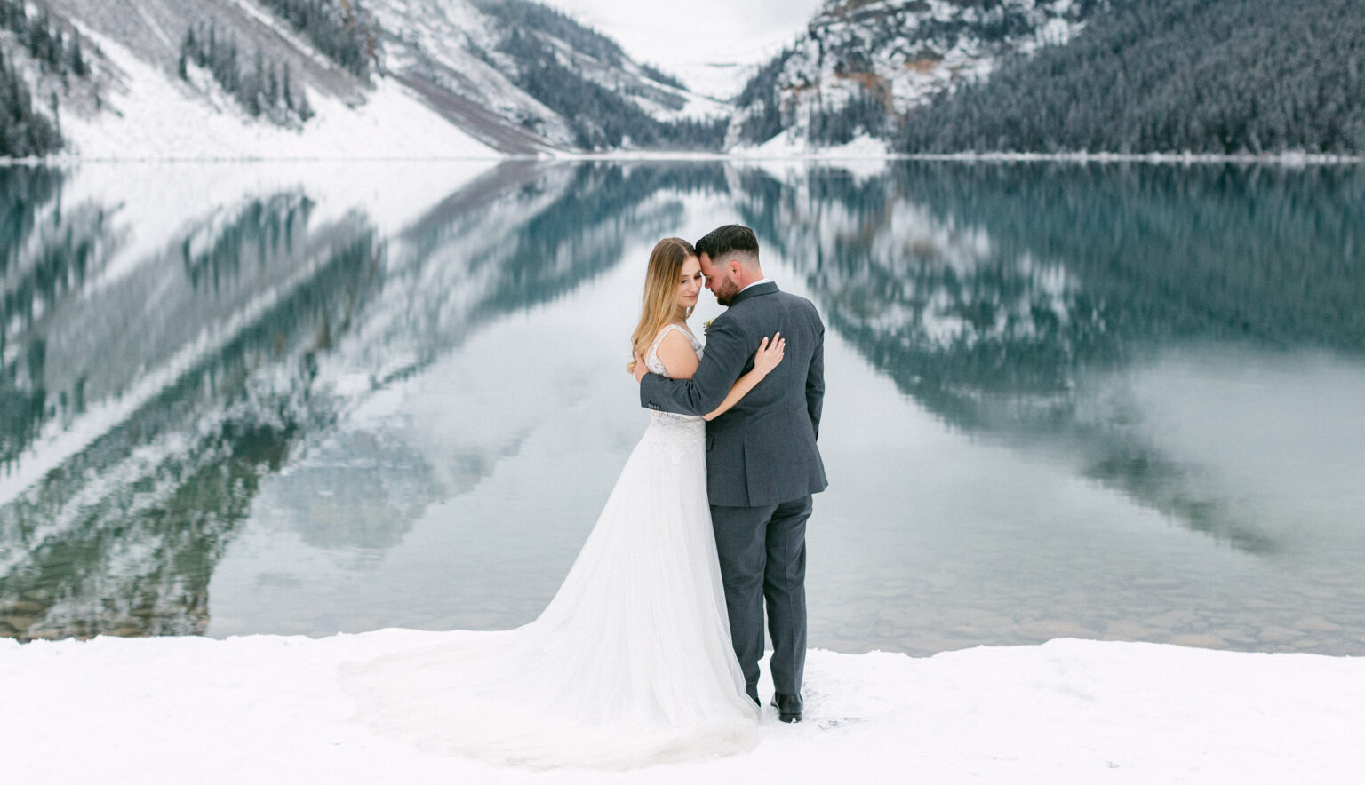 A couple embracing by a tranquil, icy lake surrounded by snow-capped mountains, capturing a romantic moment in a winter wedding setting.