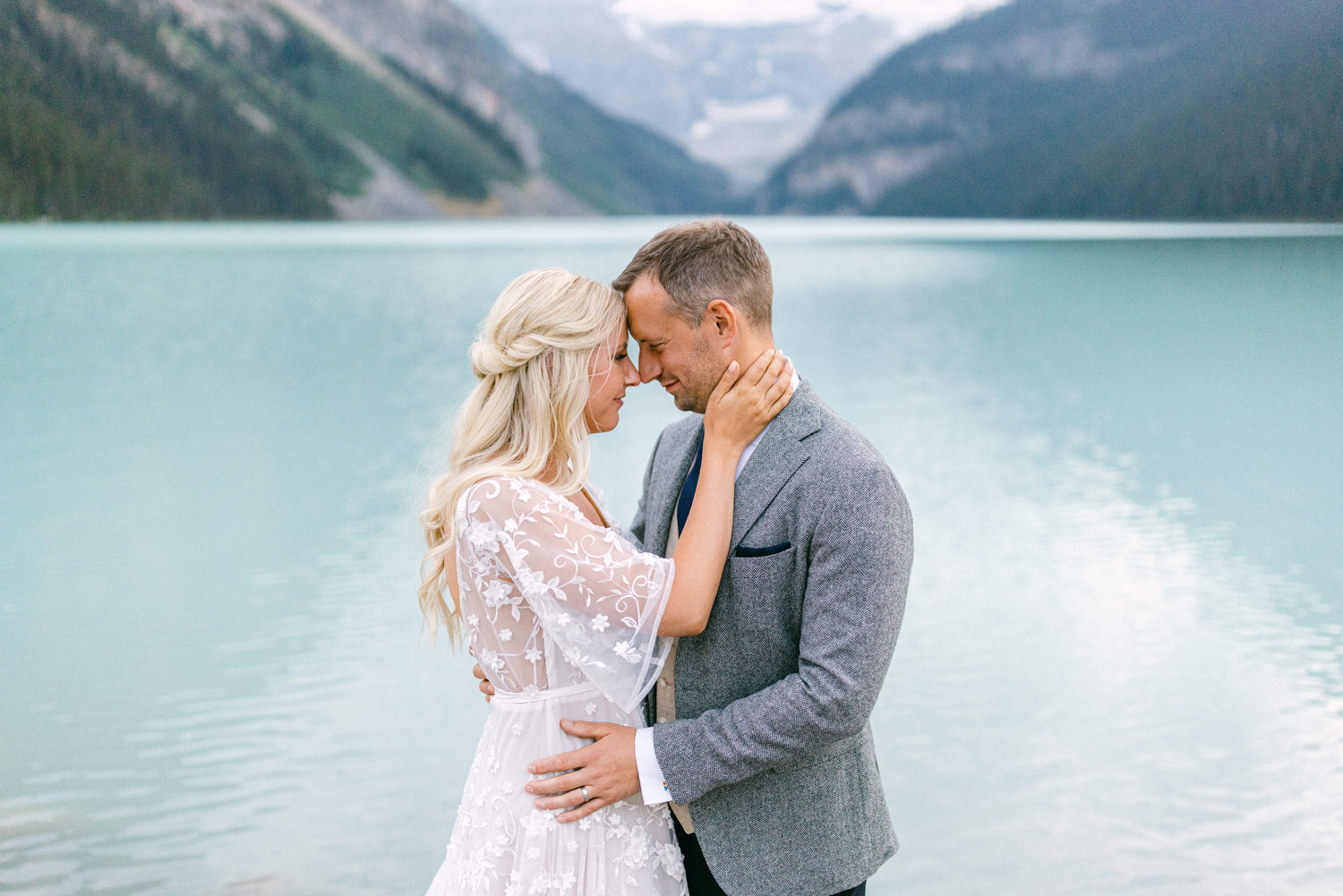 A couple gazes lovingly at each other, standing close together by the serene turquoise waters of Lake Louise, surrounded by mountains.