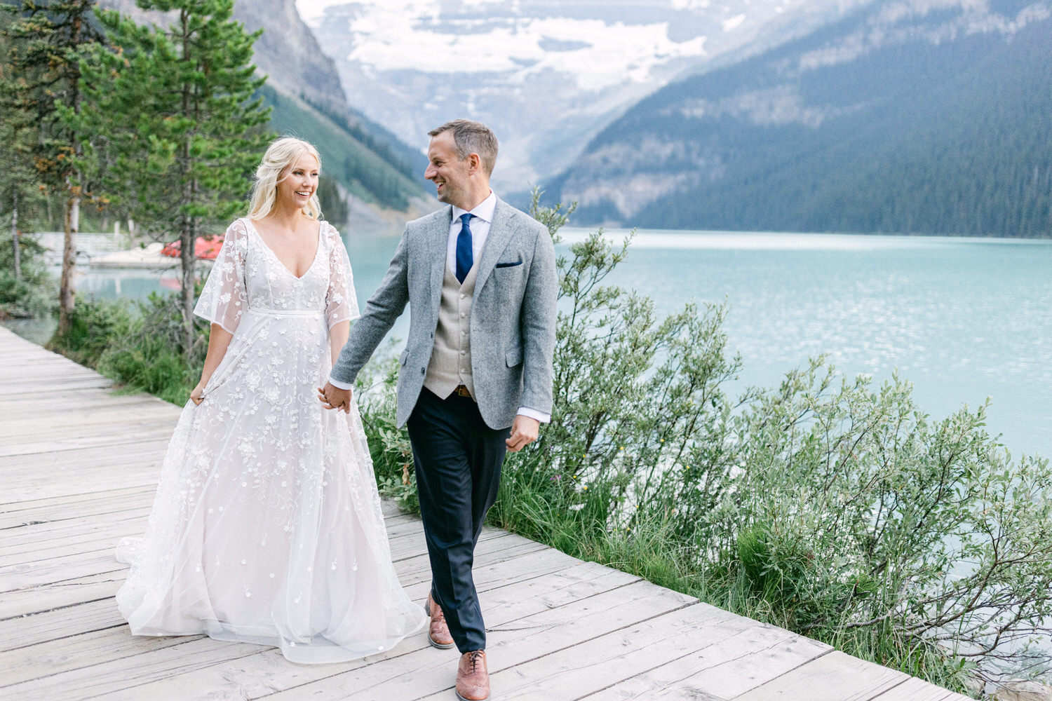 A couple walks hand-in-hand along a wooden path beside a serene turquoise lake, surrounded by lush greenery and mountains. The bride wears a delicate, embroidered white dress, while the groom is dressed in a stylish gray suit.