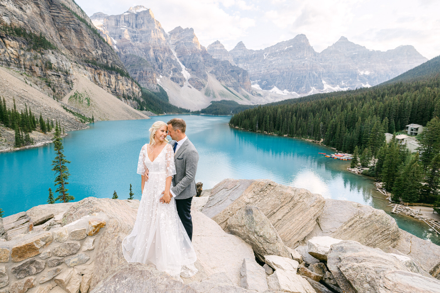 A bride and groom embrace on rocky terrain overlooking a stunning turquoise lake and towering mountains in the background.