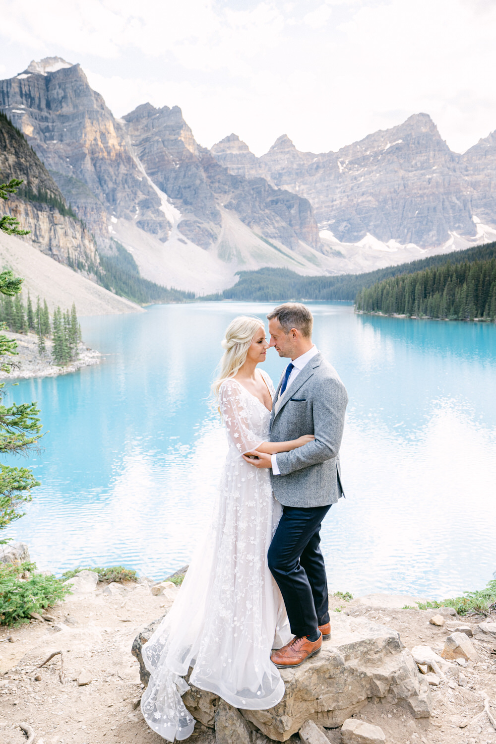 A couple stands closely together on a rock by a vibrant turquoise lake, framed by majestic mountains and lush greenery.