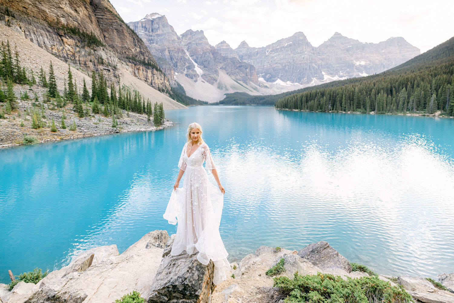 A bride in a flowing white gown stands on a rock by a stunning turquoise mountain lake surrounded by lush greenery and majestic mountains.