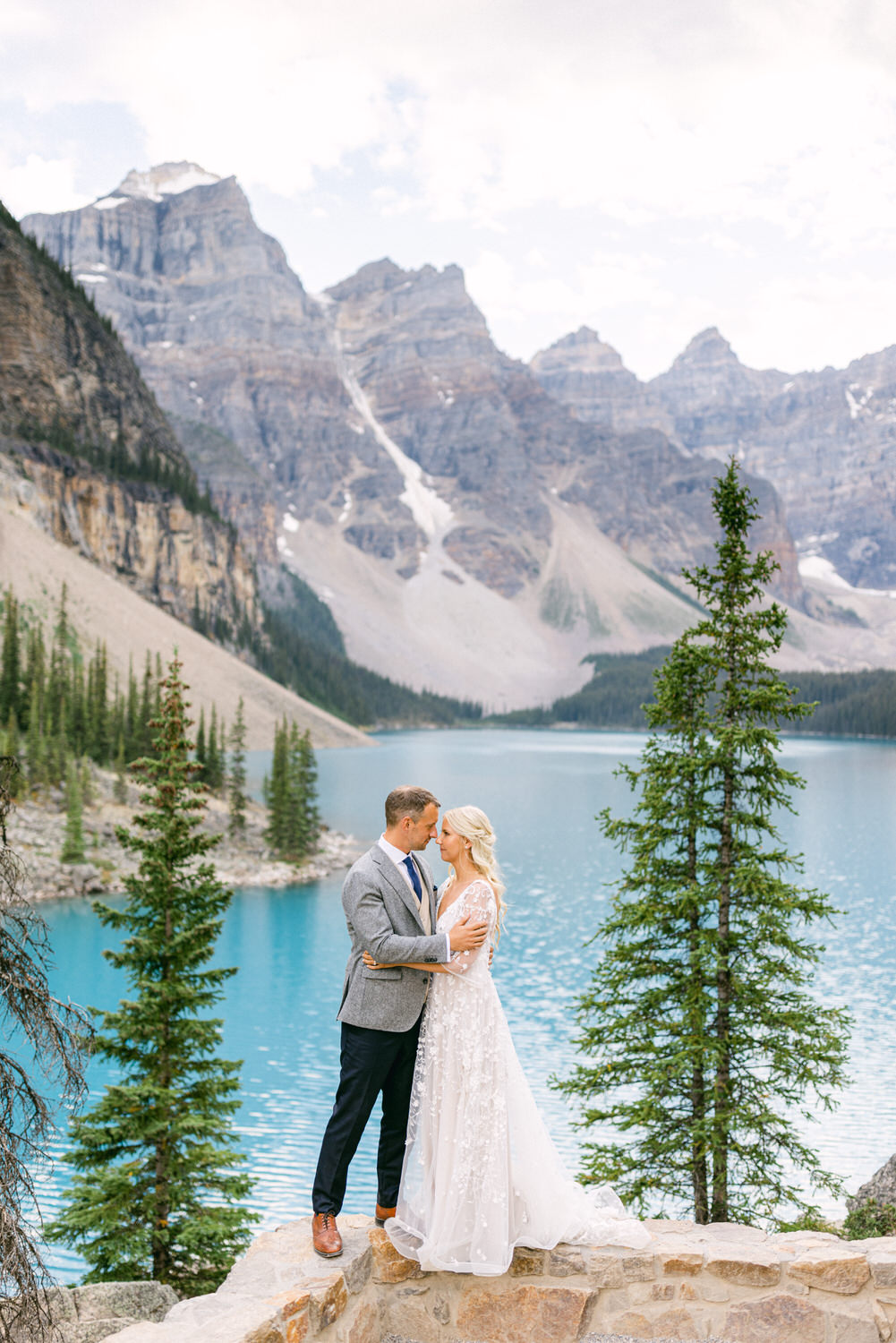 A couple embraces on a stone ledge by a vibrant turquoise lake, surrounded by towering mountains and lush greenery.