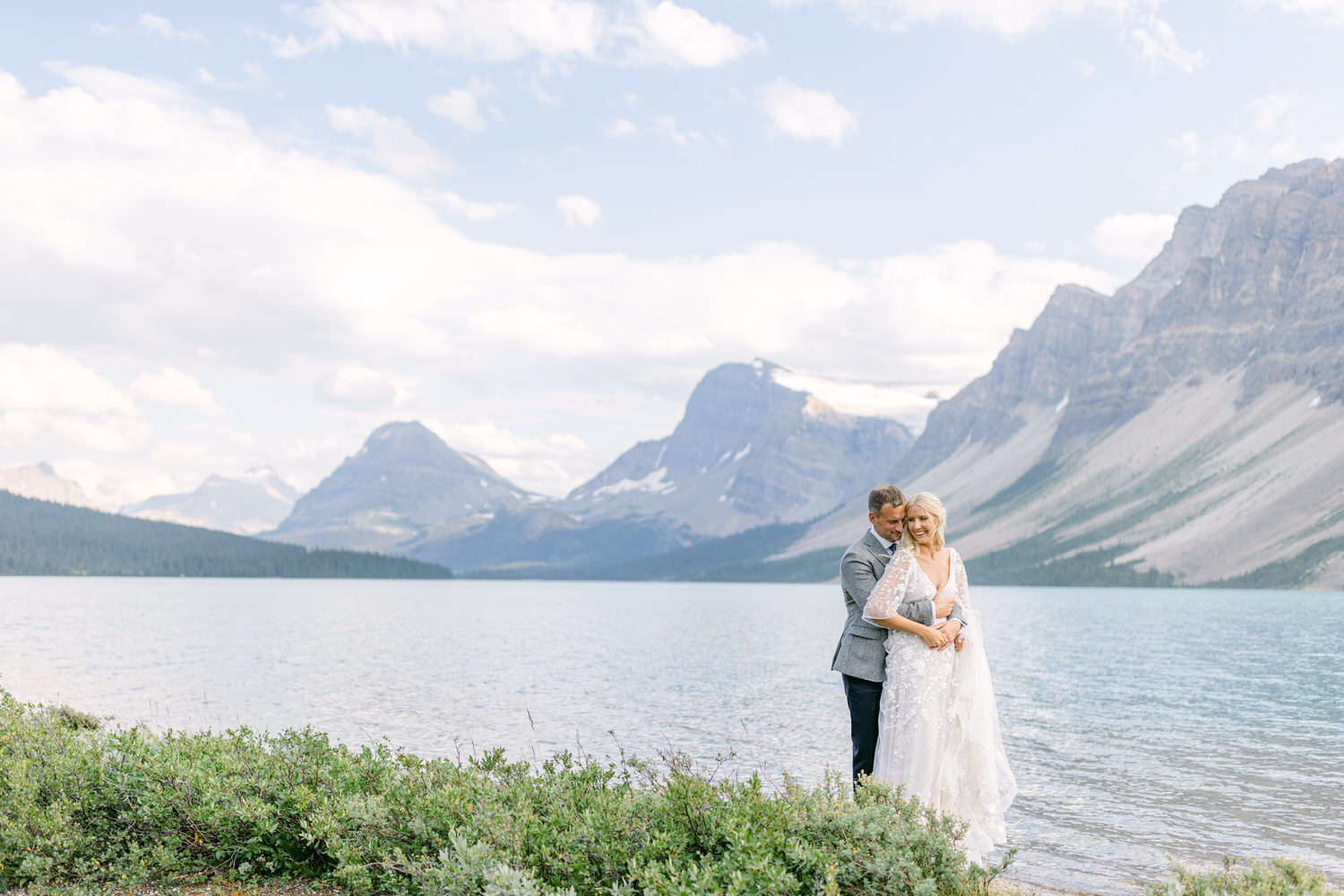 A couple embraces by a serene lake surrounded by towering mountains, capturing a moment of love in a breathtaking natural setting.