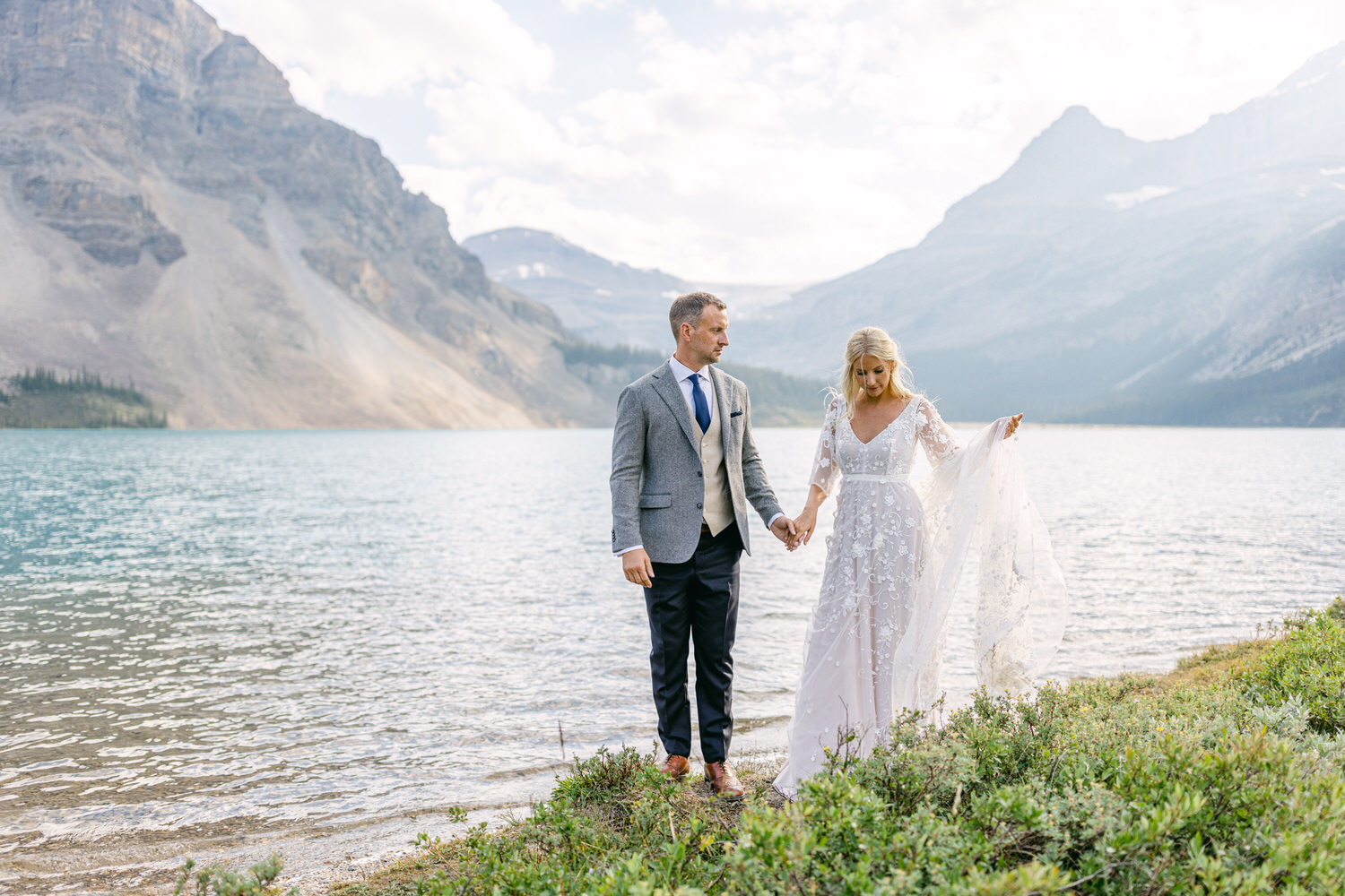 A couple holds hands by a serene lake, surrounded by mountains, showcasing a beautiful wedding setting with a stunning dress and elegant attire.