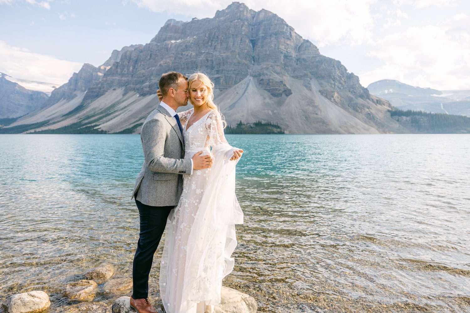 A joyful couple stands by a serene lake, surrounded by mountains, as they share a romantic moment near the water. The woman wears a flowing lace wedding dress while the man looks casual yet stylish in a blazer.