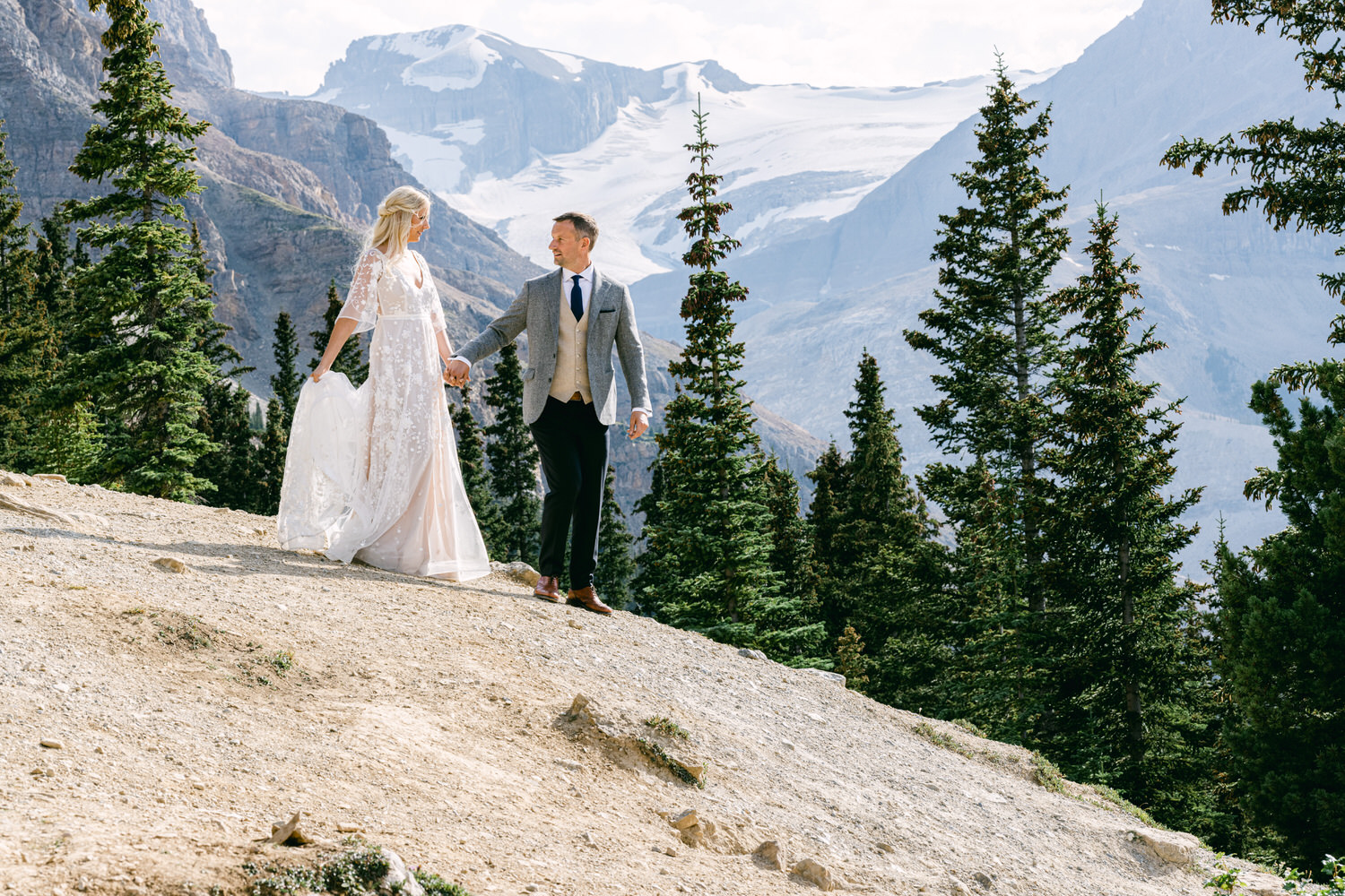 A couple walks hand-in-hand on a scenic mountain path, surrounded by lush greenery and a stunning glacier backdrop.