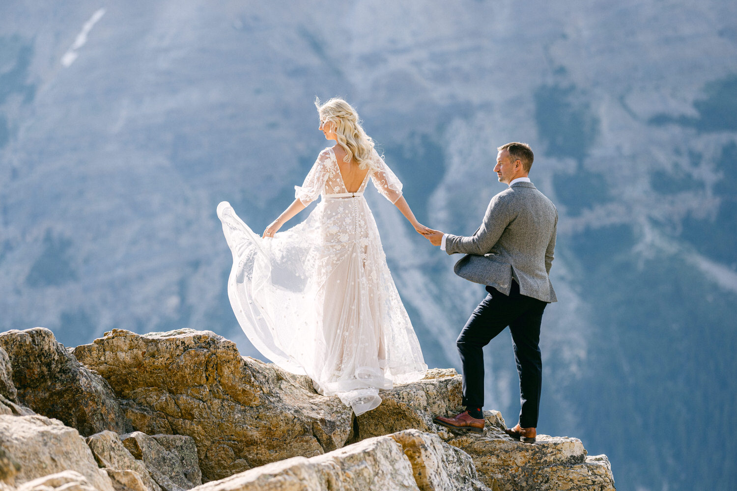 A couple embraces their love on a rocky ledge, with the bride in a flowing gown and the groom in a stylish suit, set against a breathtaking mountain backdrop.