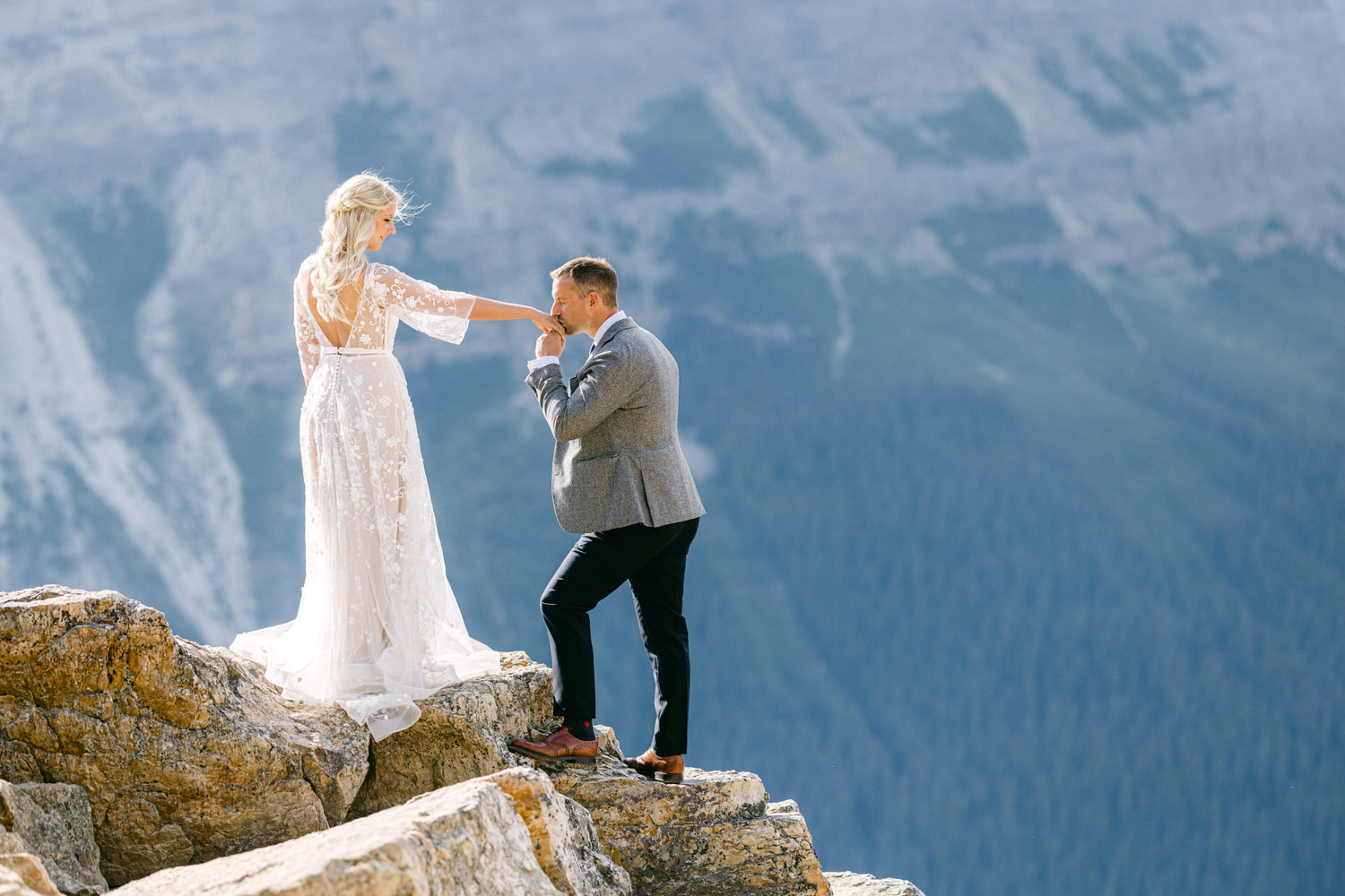 A couple shares a special moment on a rocky ledge, with the man kissing the woman's hand against a scenic mountain backdrop.