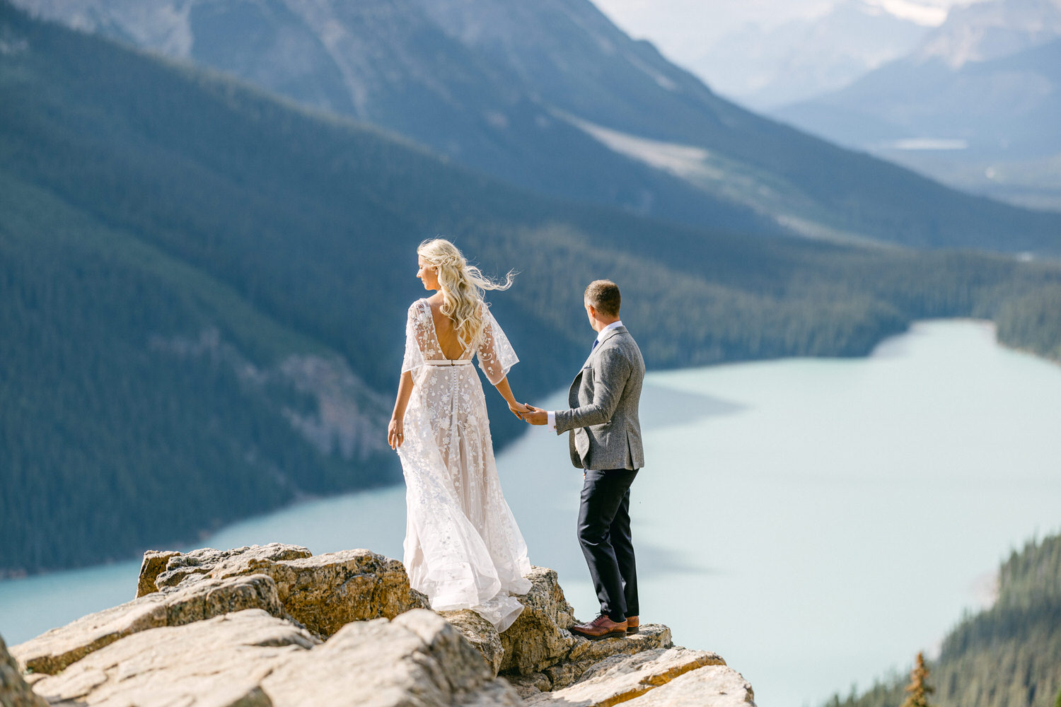 A couple stands hand-in-hand on a rocky outcrop overlooking a stunning mountain lake, surrounded by lush greenery and majestic peaks. The woman wears a flowing white wedding dress, while the man is dressed in a smart gray jacket and dark pants.