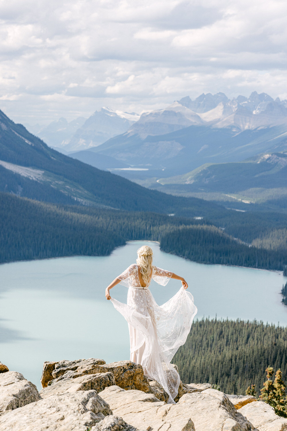 A woman in a flowing white dress stands on rocky terrain, gazing at a breathtaking mountain landscape with a serene lake and lush forests in the background.