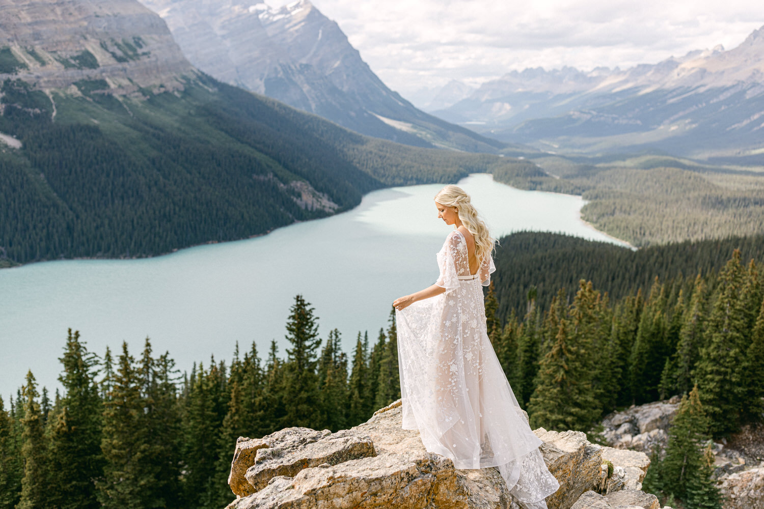 A bride in a flowing lace gown stands on a rocky outcrop overlooking a serene lake surrounded by forests and mountains.