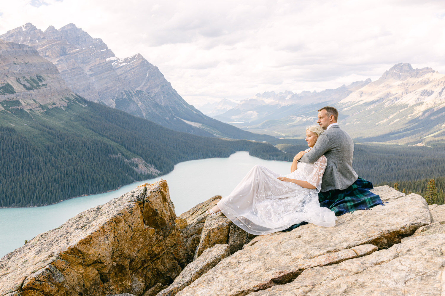 A couple embraces on a rocky overlook, with a stunning mountain and forest backdrop, showcasing love amidst nature's beauty.