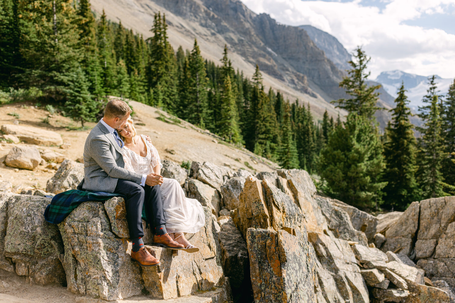 A couple shares an intimate moment on a rocky outcrop in a mountainous landscape, surrounded by pine trees and stunning vistas.