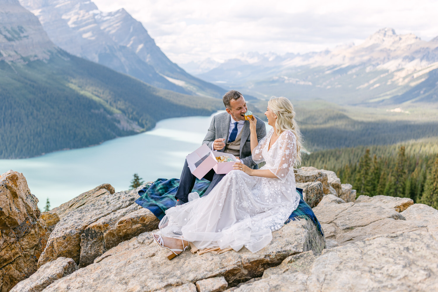 A couple enjoys a serene picnic on a rocky ledge with a breathtaking lake and mountain backdrop, showcasing love and celebration.