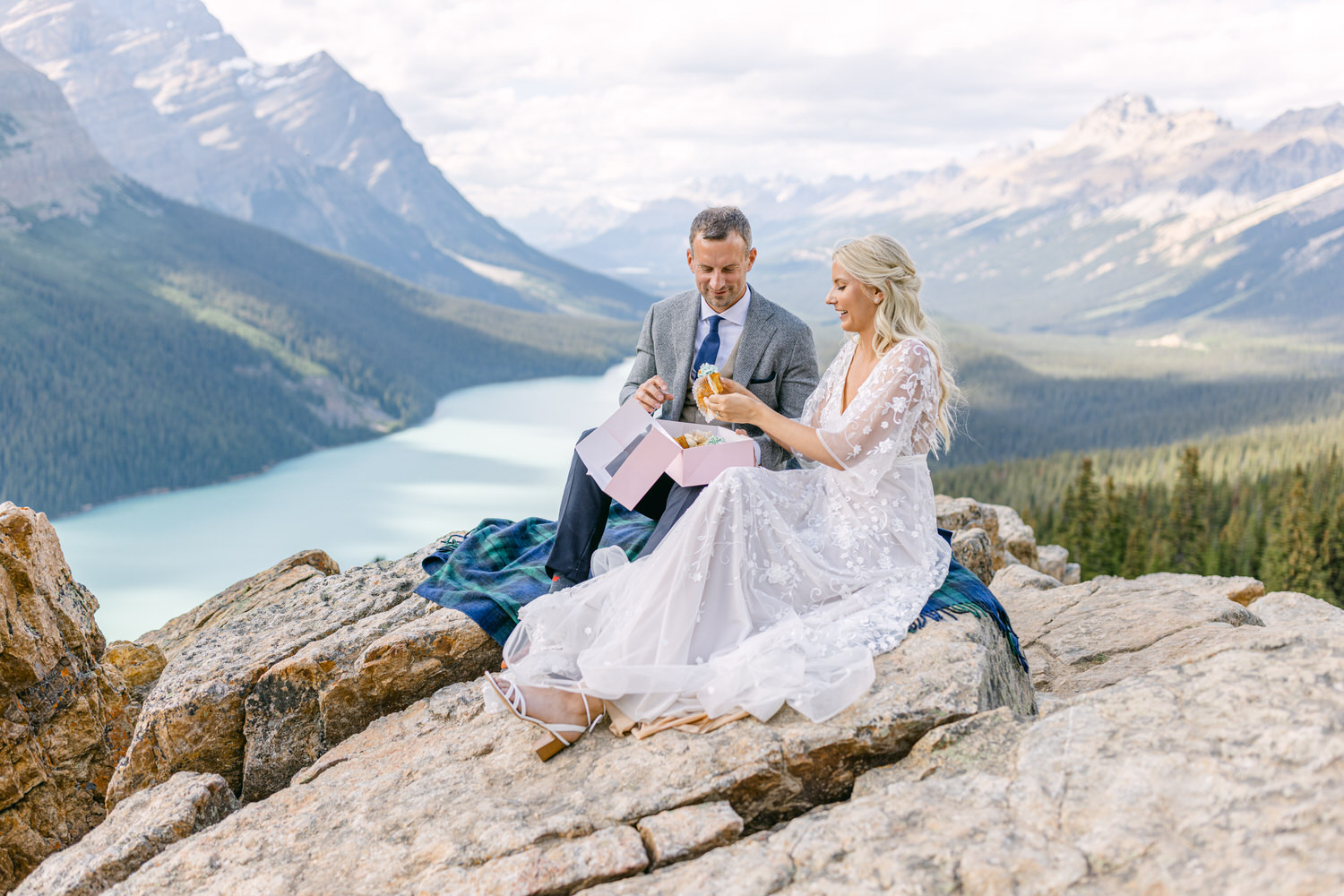 Couple enjoying a moment together on a rocky ledge with a breathtaking view of a lake and mountains, sharing a dessert from a pink box.