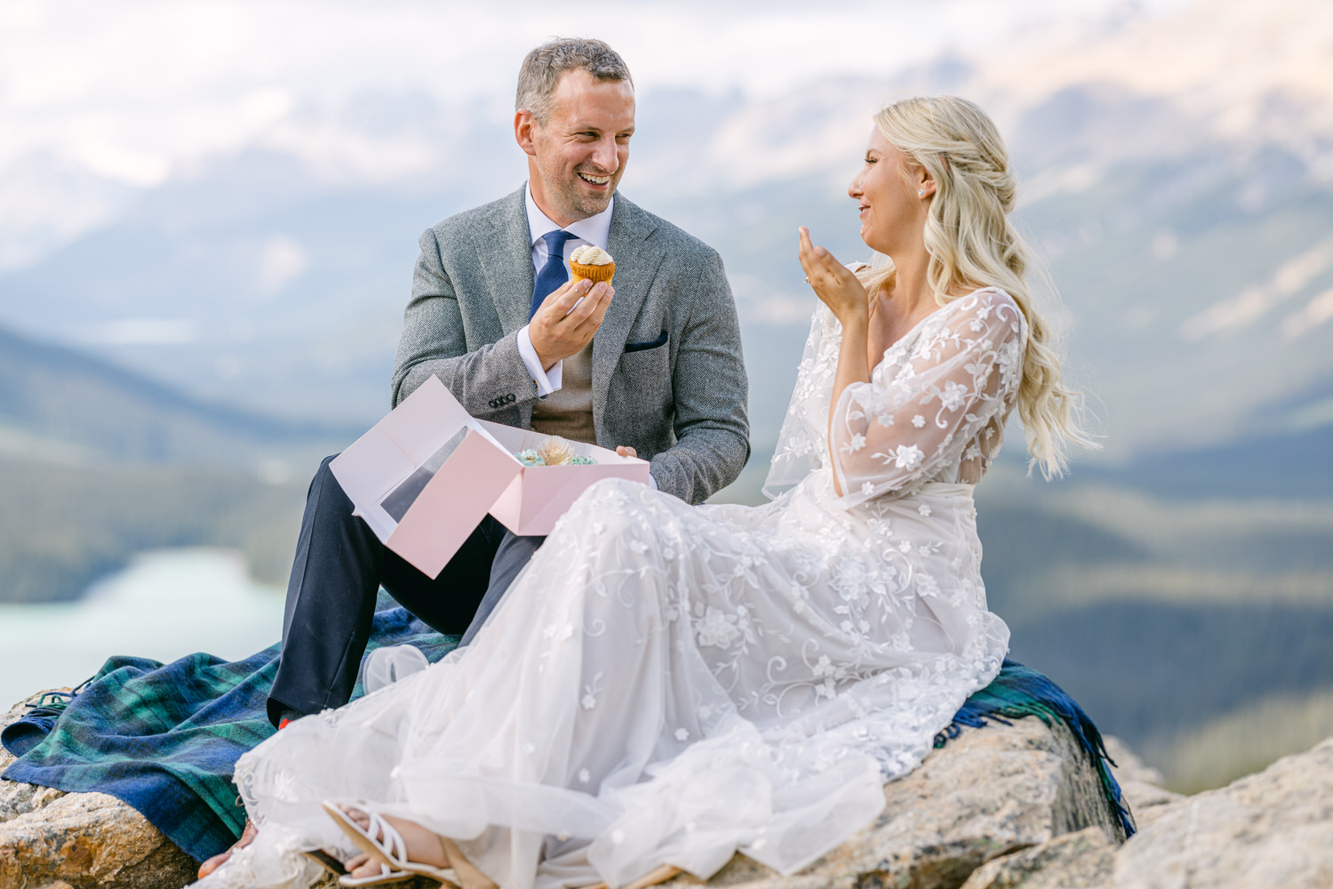 A happy couple seated on a rocky ledge in the mountains, sharing cupcakes and laughter, dressed elegantly, surrounded by breathtaking scenery.