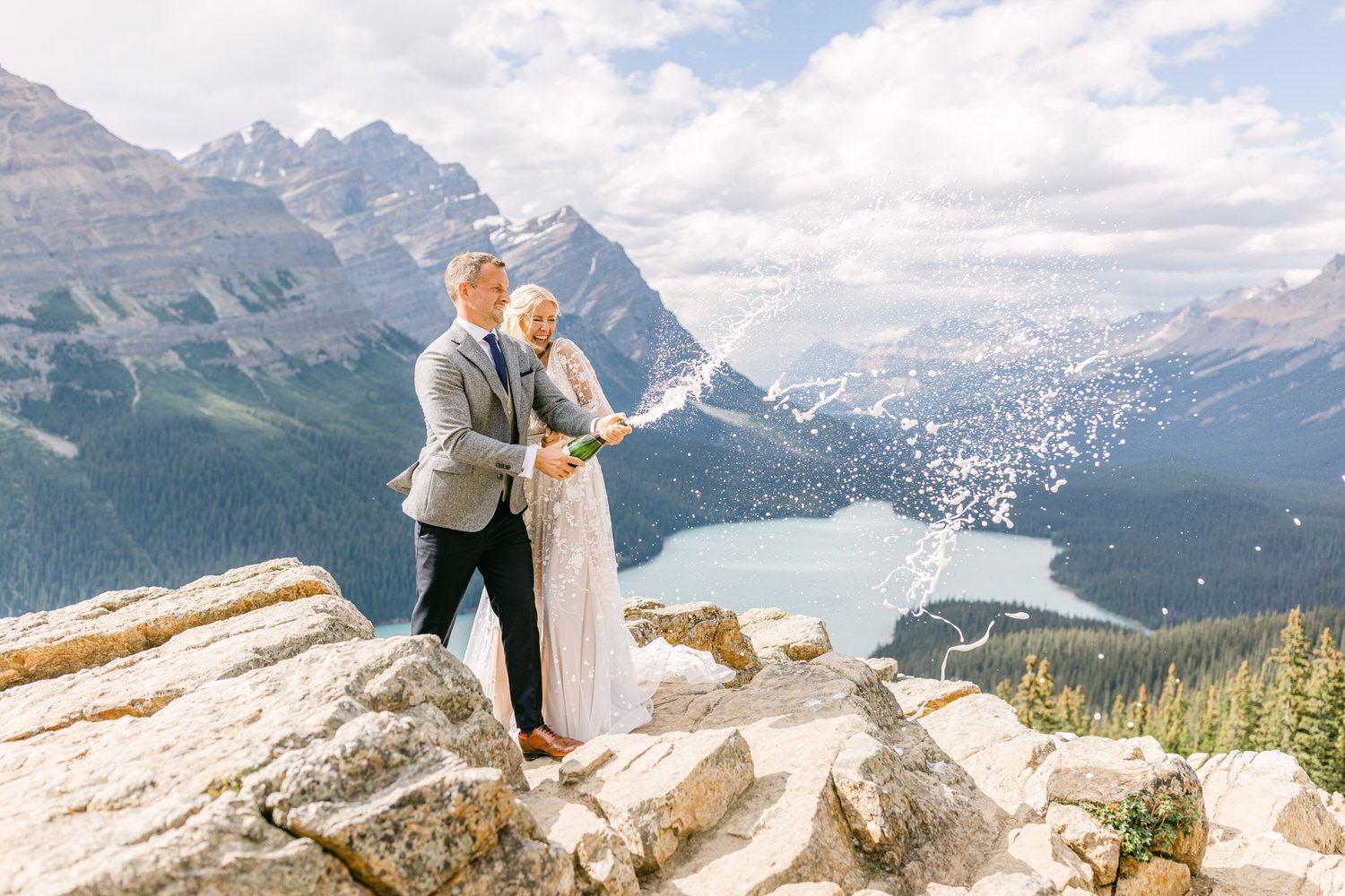 A joyful couple pops champagne on a rocky mountain peak, celebrating against a stunning landscape of mountains and a turquoise lake in the background.