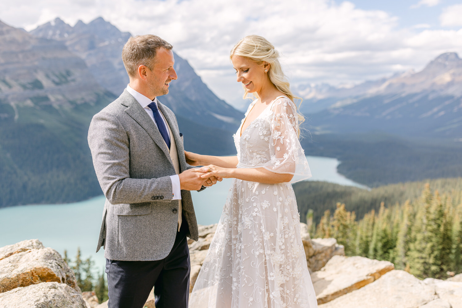 A couple exchanging rings during an outdoor wedding ceremony with a mountainous landscape and lake in the background.