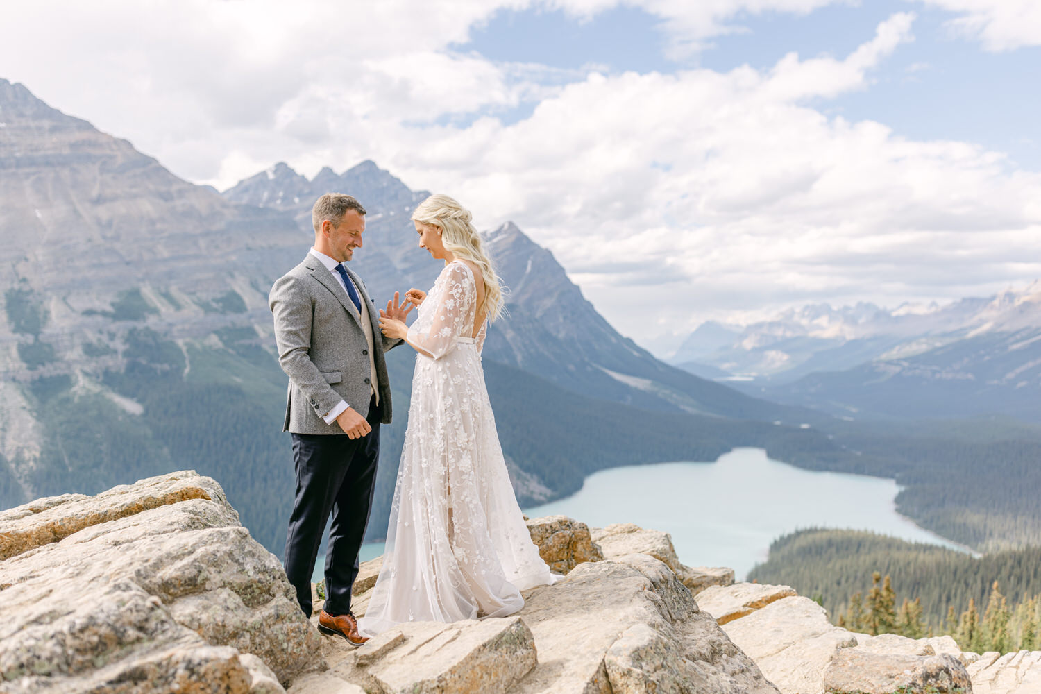 A couple exchanges vows on a rocky ledge with a stunning mountain and lake backdrop, showcasing their love in a picturesque outdoor setting.