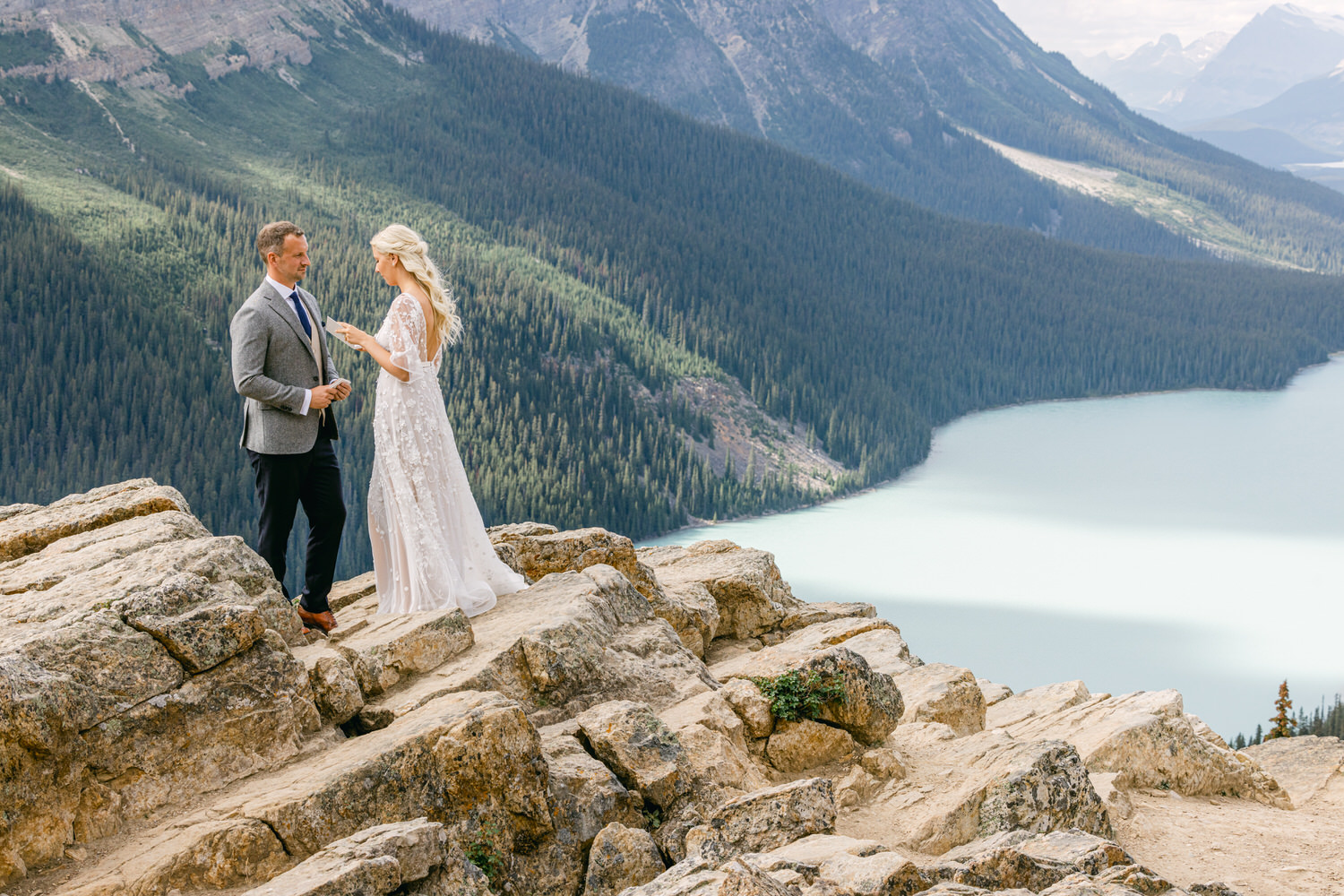 A couple exchanging vows on a rocky outcrop with a scenic view of mountains and a lake in the background.