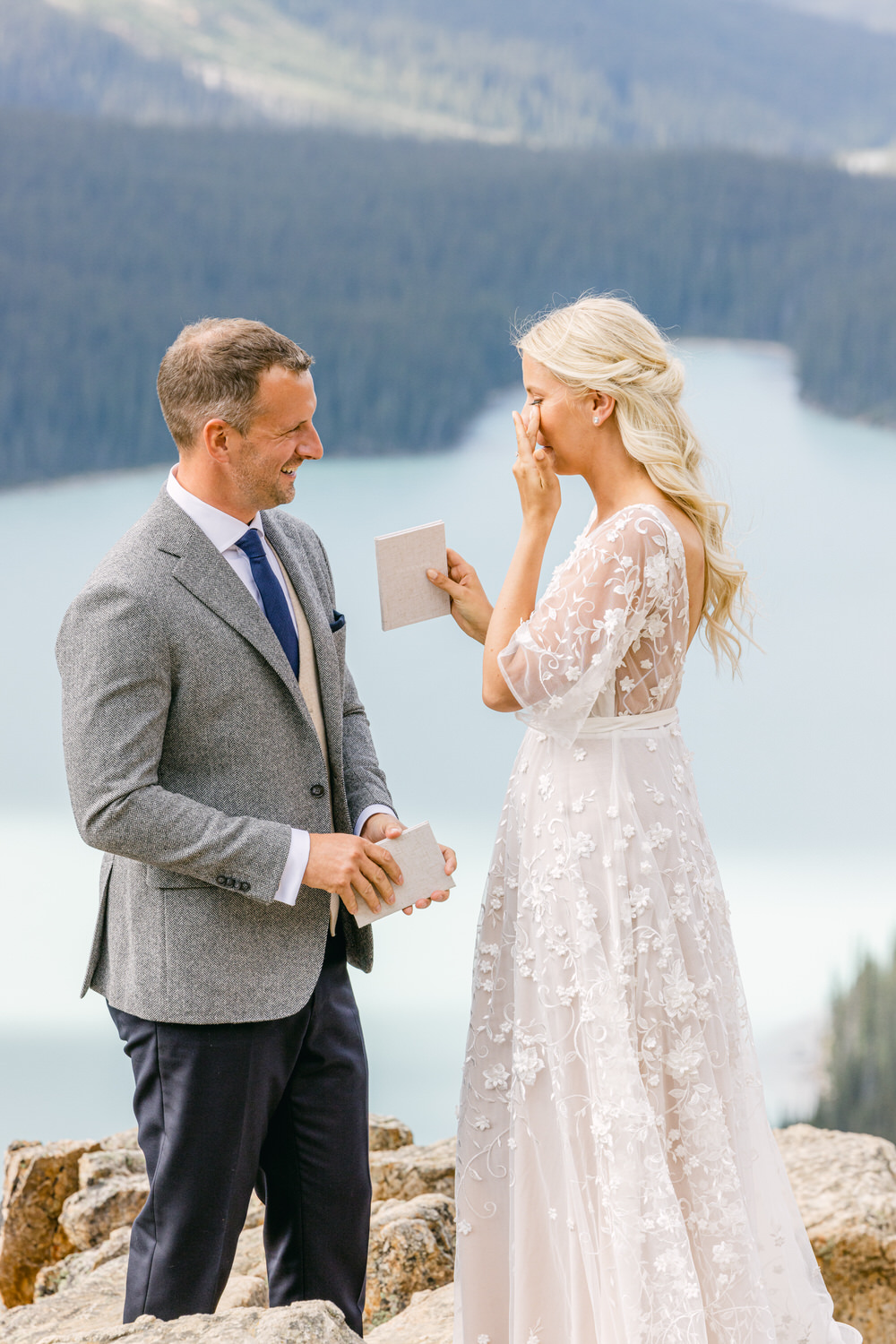 A couple exchanging heartfelt vows on a rocky ledge with a breathtaking mountain lake backdrop. The bride, in a floral lace gown, wipes a tear of joy while the groom smiles warmly, holding a small booklet.