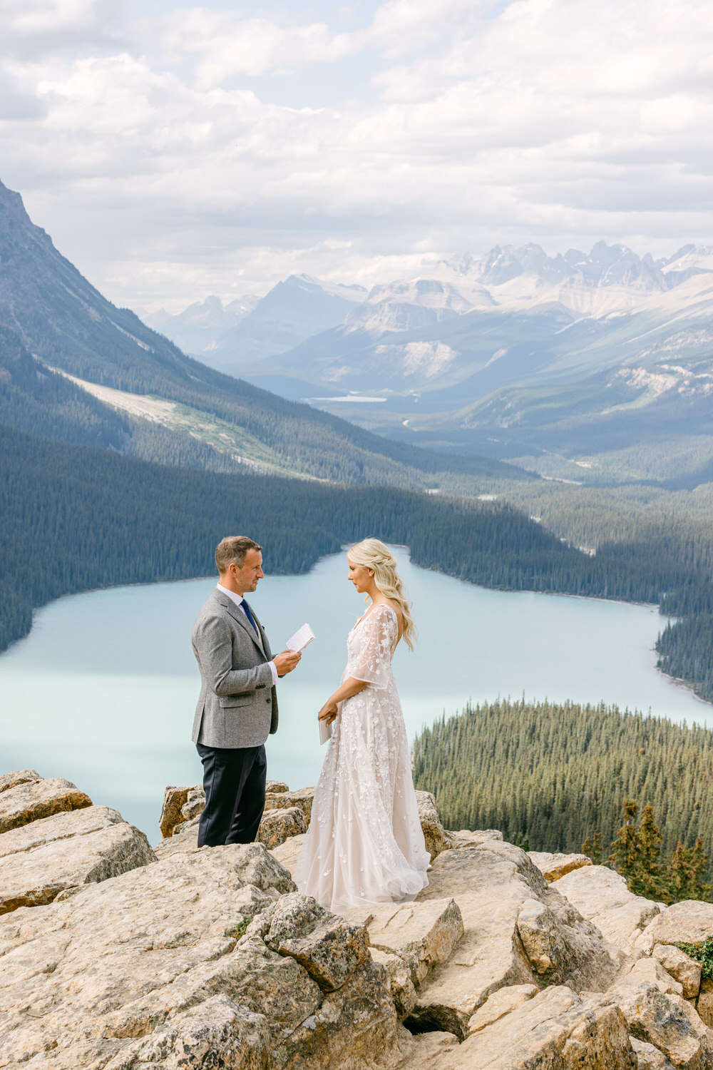 A couple exchanging vows atop a rocky overlook with a stunning lake and mountains in the background.