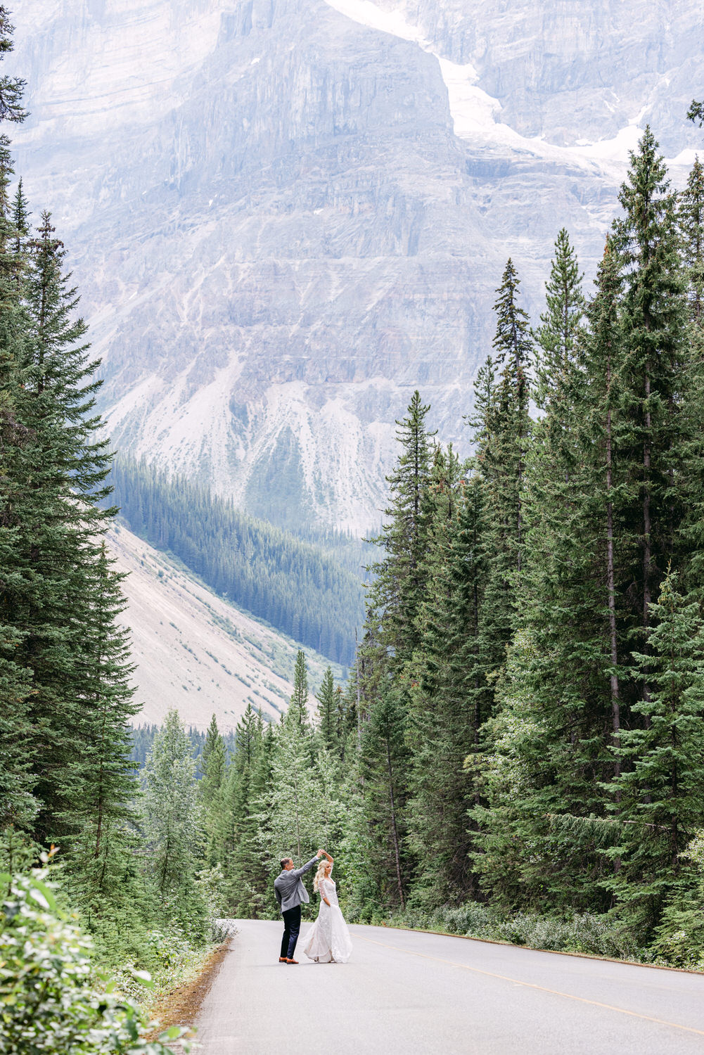 A couple dances joyfully on a serene mountain road surrounded by lush greenery and towering peaks.